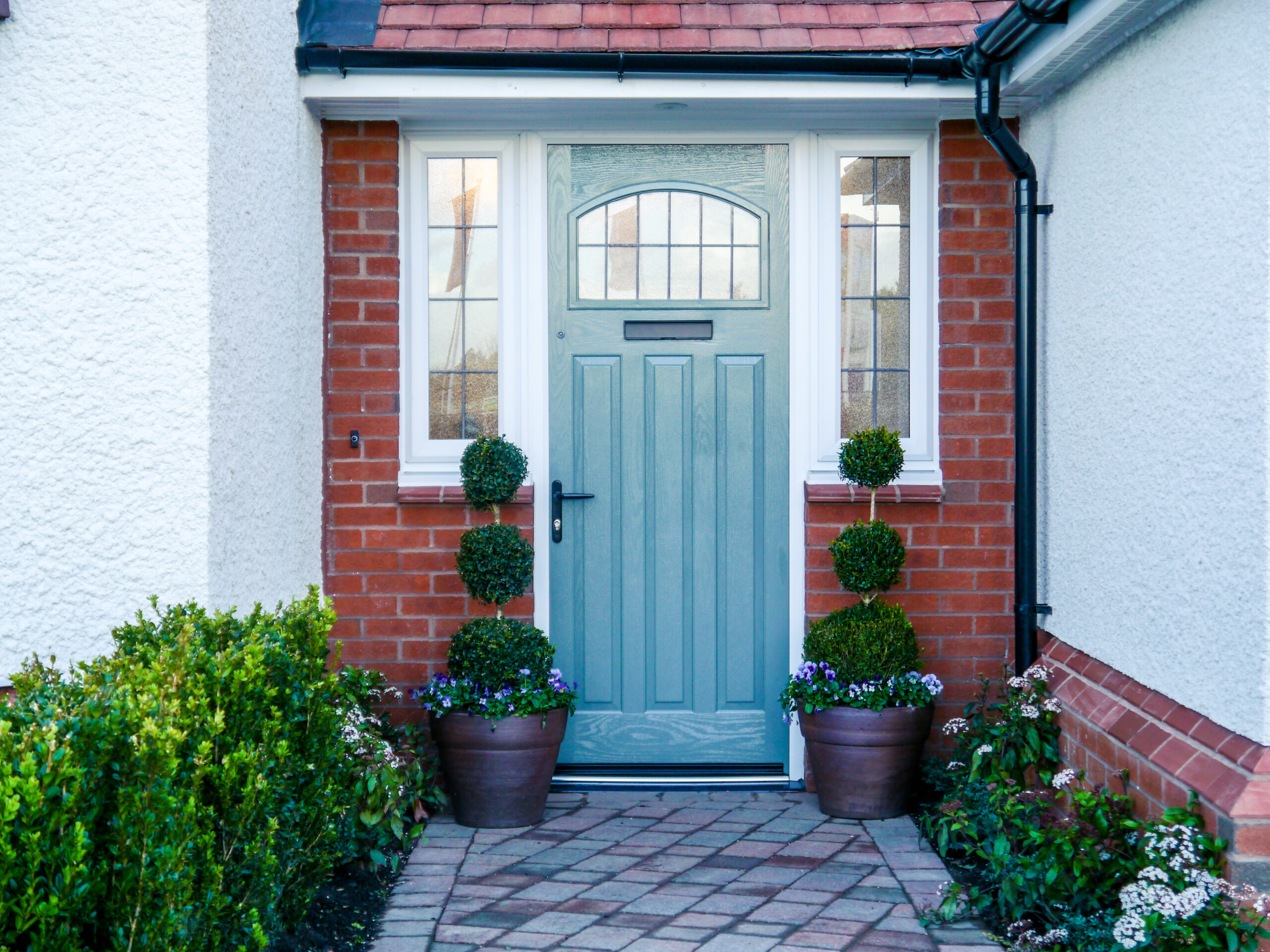 Painted blue front door with arched window and black handle, flanked by stained-glass sidelights and two topiary pots.