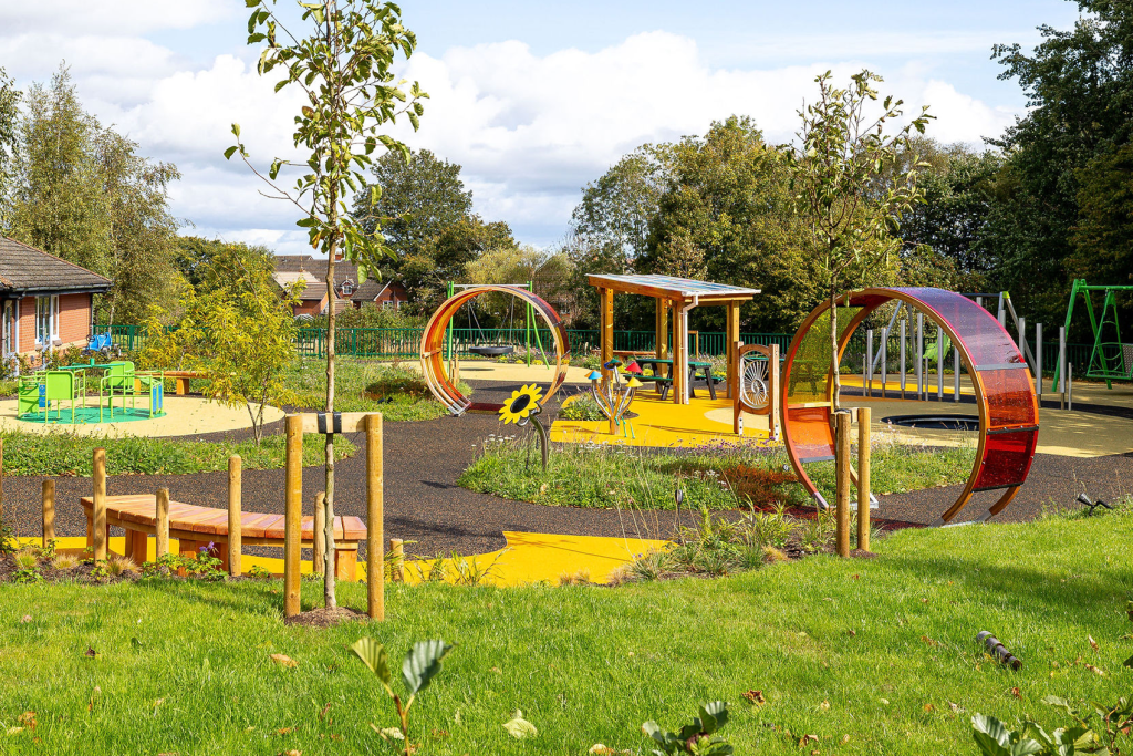 Colourful modern children's playground with circular climbing tunnels, benches, picnic shelter and grassy lawns under trees