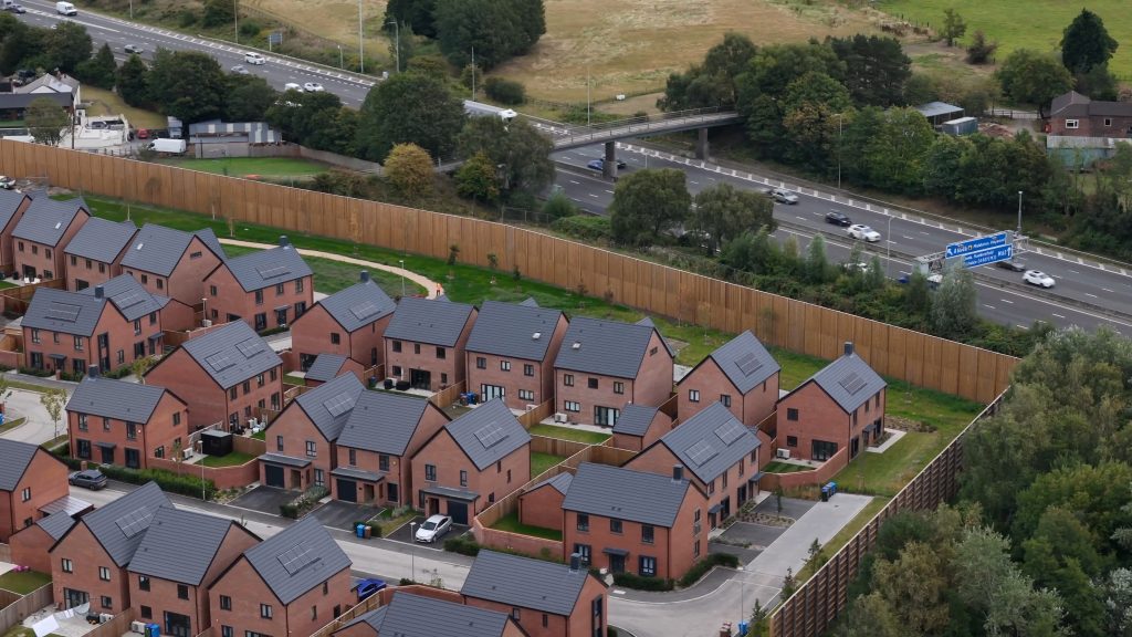Aerial view of newly completed brick houses within a residential development, bordered by a tall timber noise barrier.